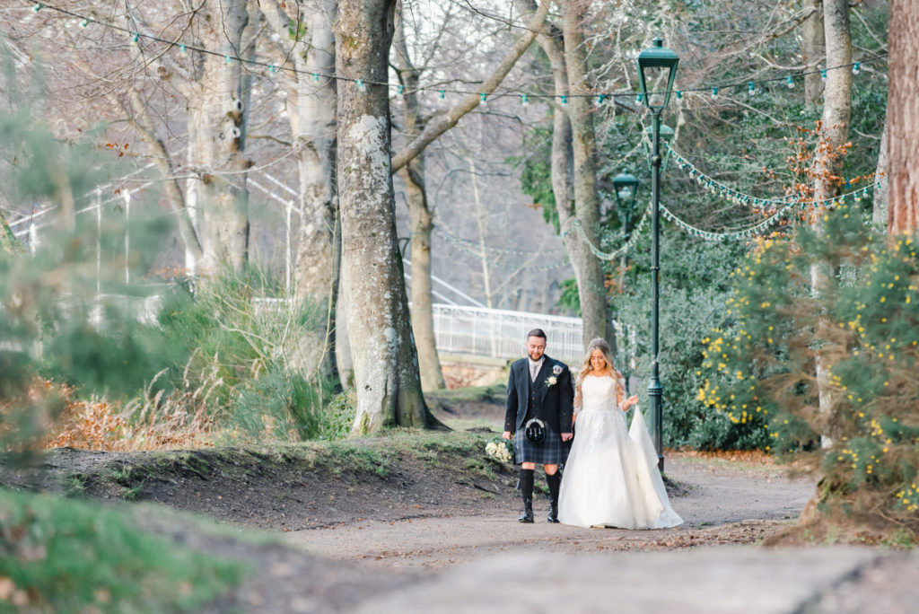 A Caucasian bride and groom holding hands and walking on a woodland path in front of a lamppost, tall trees and a footbridge