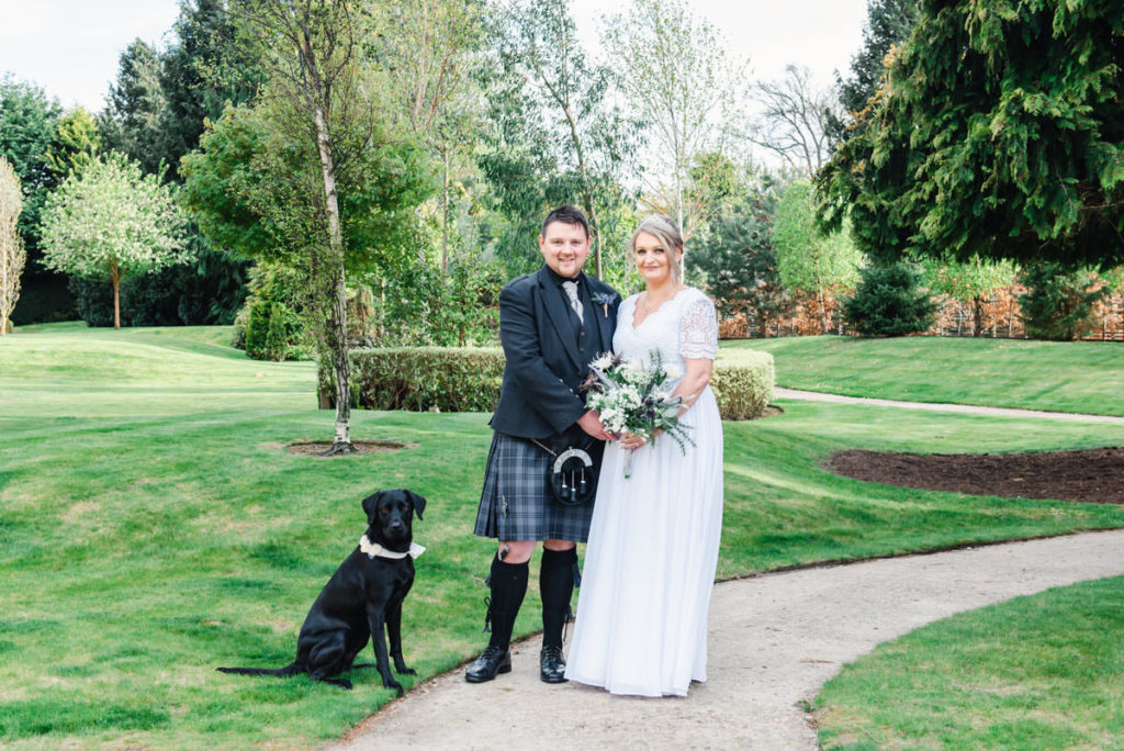 A black dog sitting on grass and a bride and groom holding flowers standing on a path in a garden with trees and shrubs