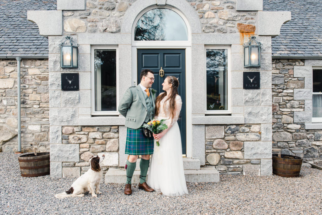 A white and brown dog looking up at a bride and groom facing each other outside the black door or a smart stone building