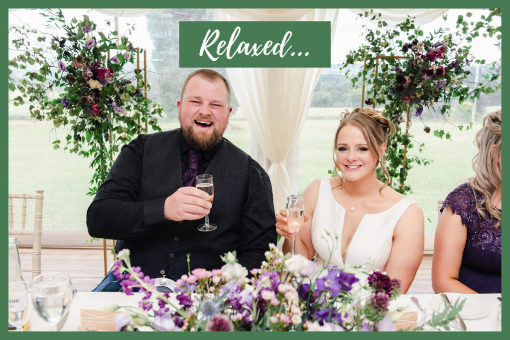 A Caucasian groom and bride holding glasses of champagne sitting at a table, smiling, in a marquee with purple flower decorations