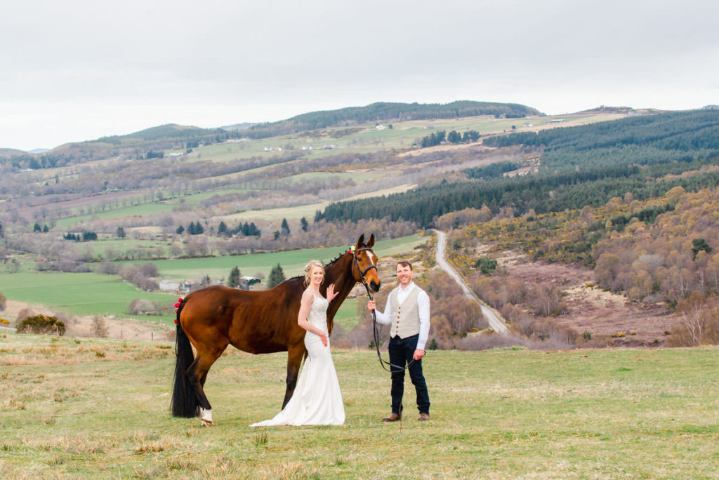 A bride in a white dress and a groom in trousers and waistcoat holding a brown horse on grass with hills in the background