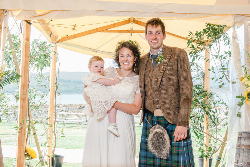 A bride in a white dress holding a baby girl standing beside a groom in a kilt in a marquee entrance with grass beyond