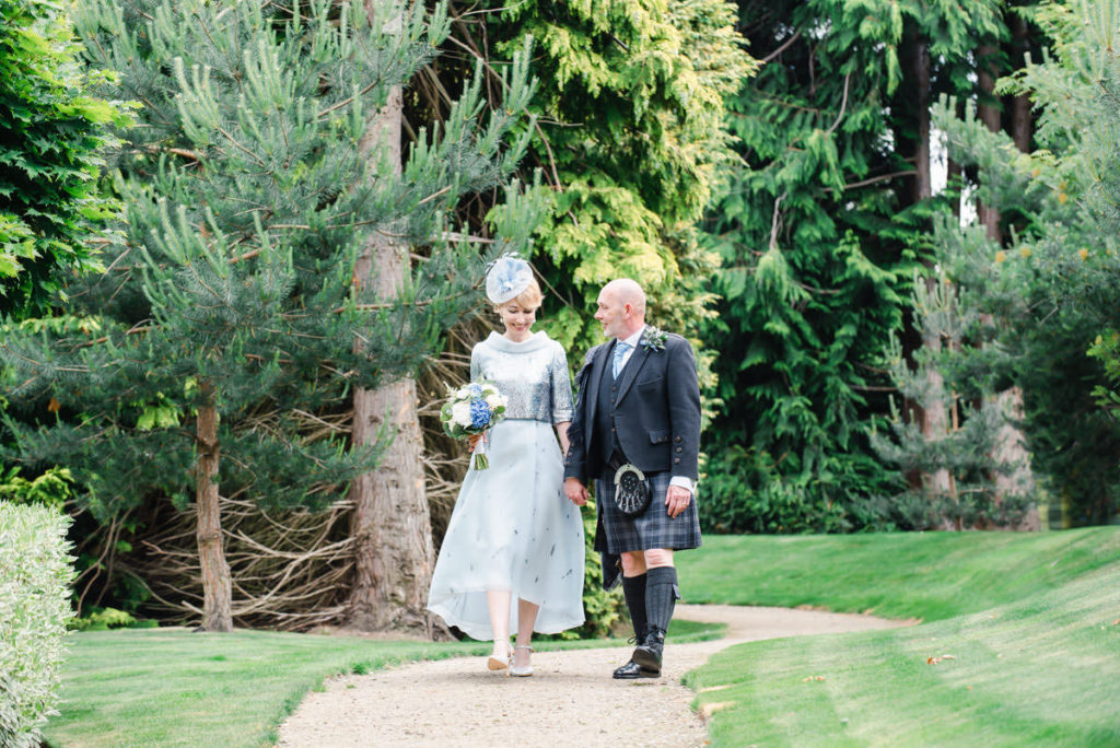 A man in a kilt and a woman in a blue dress and hat holding hands and walking on a stone path beside grass in front of trees