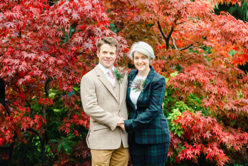 White man and woman, both dressed in suits, holding hands and standing in front of red foliage
