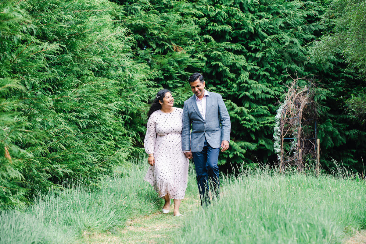 An Indian couple in a pink dress and a grey suit holding hands and walking on a path through long grass in front of a hedge