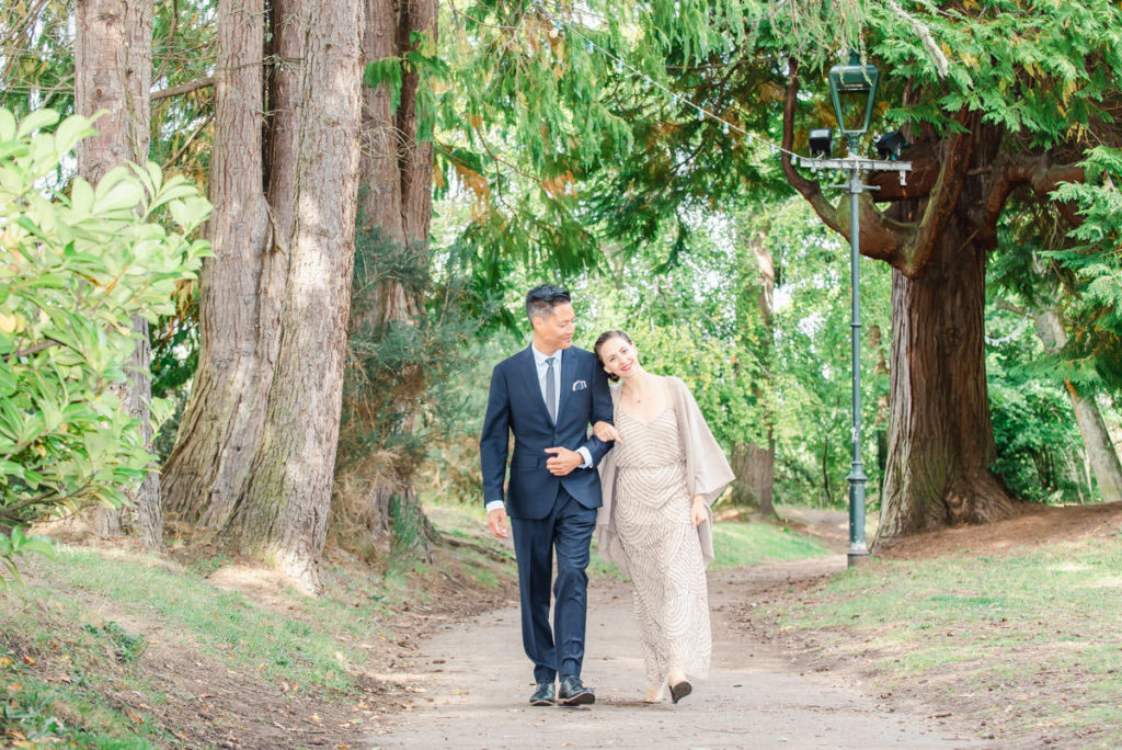A Caucasian woman walking with her head on the shoulder of an Asian man on a woodland path in front of trees and a lamppost
