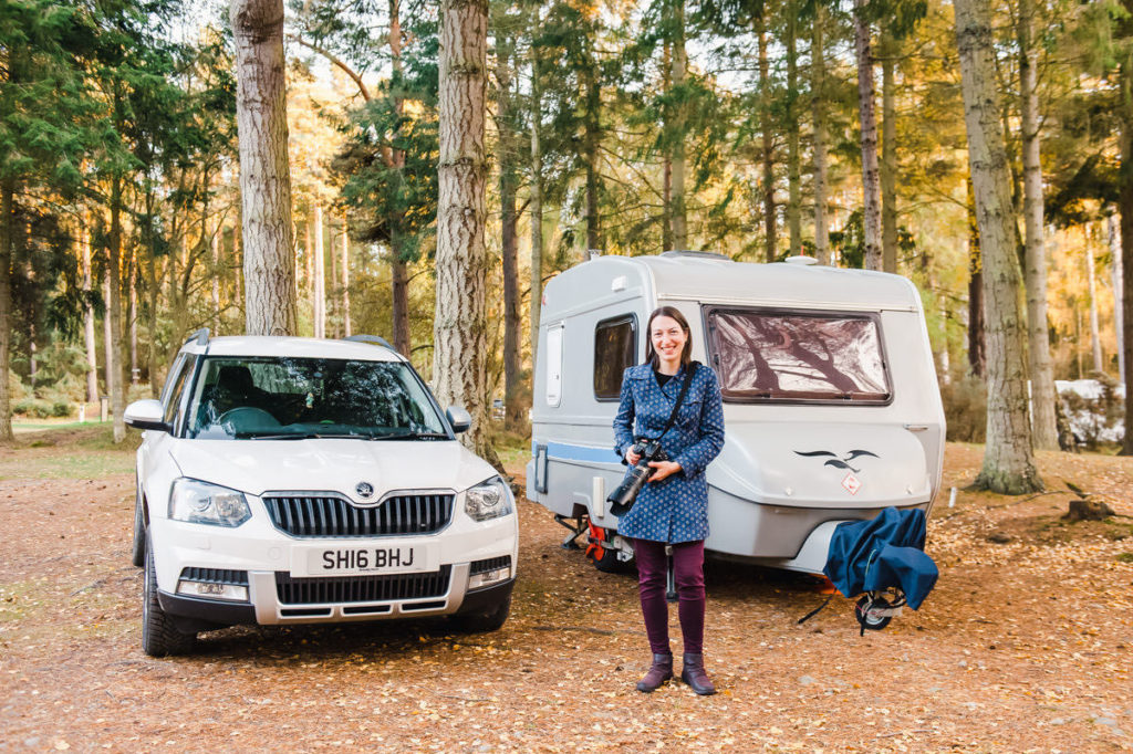 A woman holding a camera standing in front of a white car and small grey caravan in a woodland with tall trees
