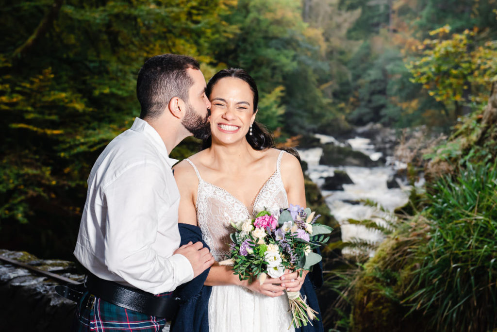 Latin American bride holding flowers and grinning while being kissed on the cheek by her groom in front of a river and trees