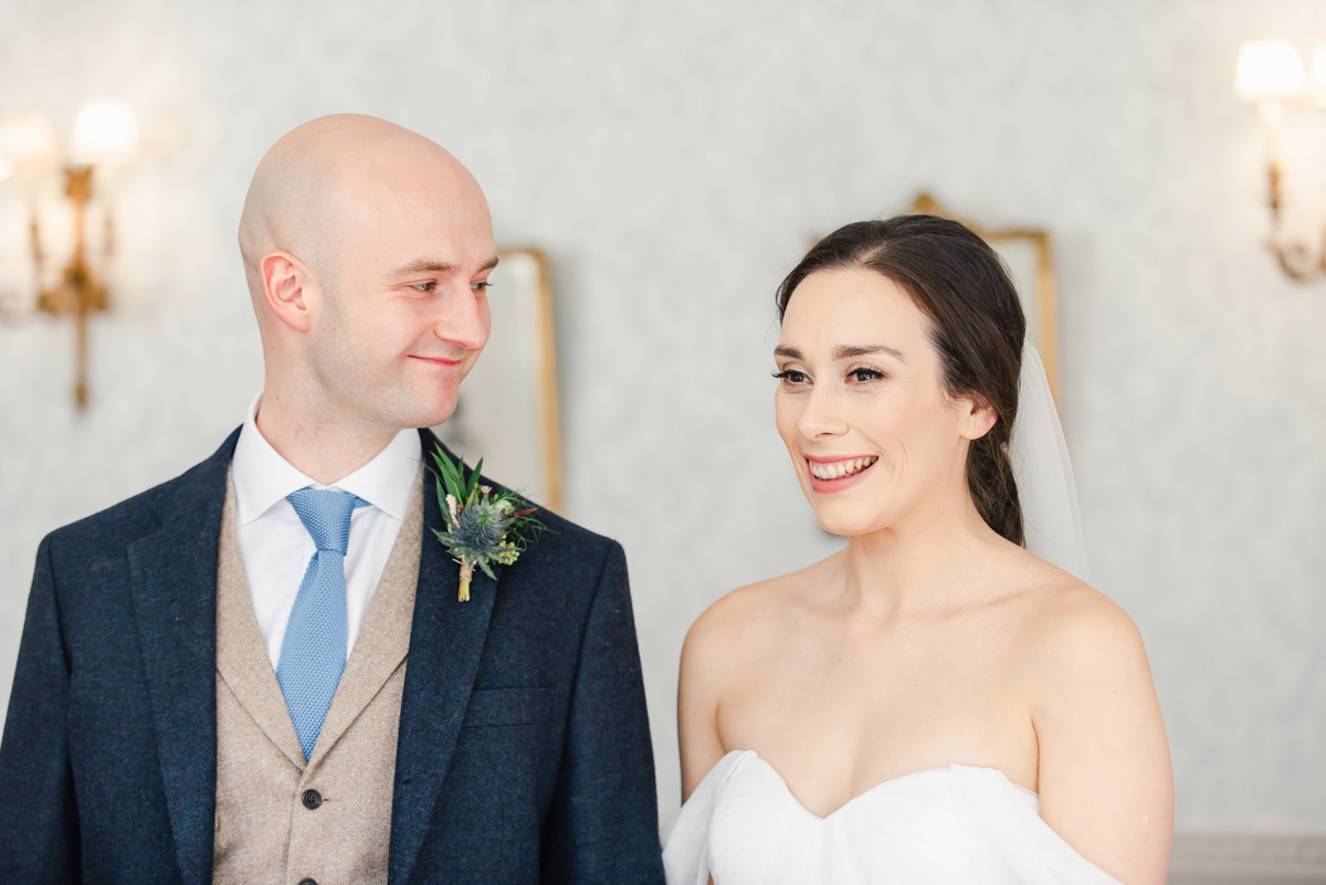 A groom looking lovingly at his smiling bride in a room with cream walls