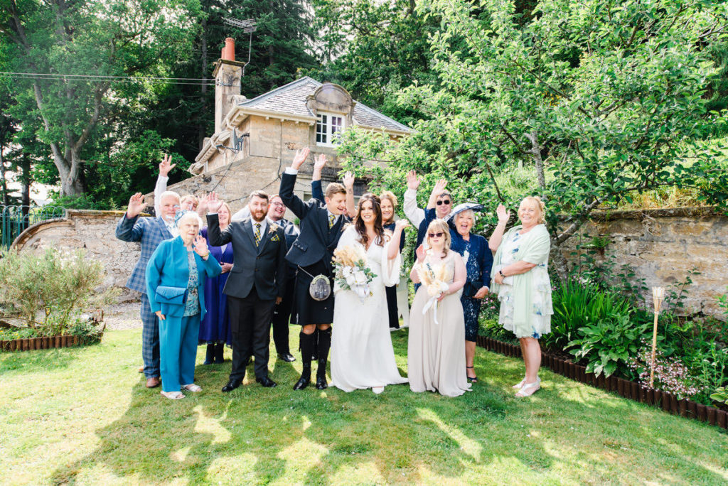 A small group of wedding guests with a bride and groom smiling and waving on a lawn in front of a two-storey stone house