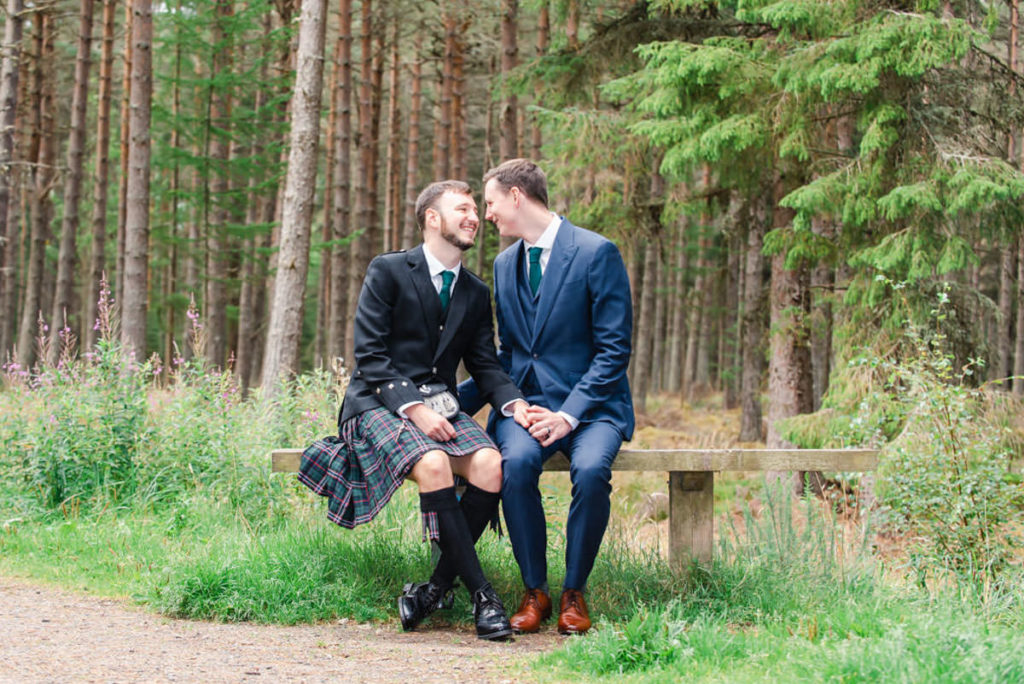 A groom in a kilt and a groom in a suit smiling at each other and sitting on a wooden bench in front of trees