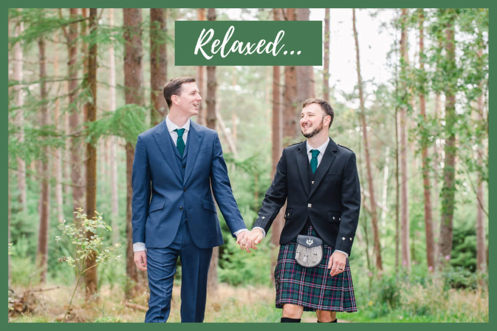 Two white men, one wearing a suit and one wearing a kilt, holding hands and smiling while walking through a woodland