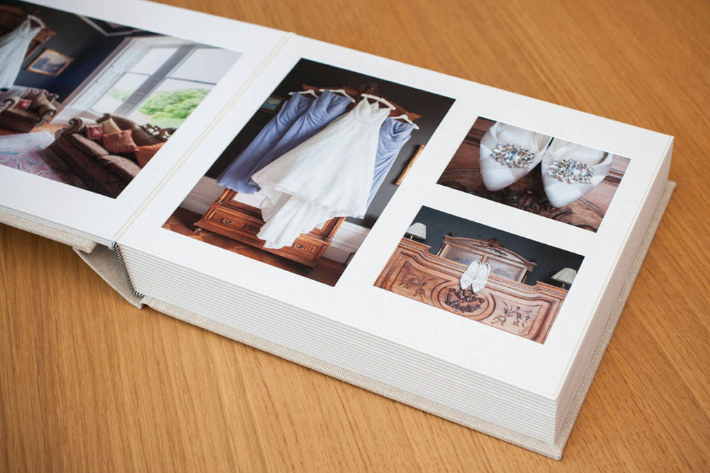 A wedding album on a wooden surface open at a double page spread showing a white wedding dress and shoes