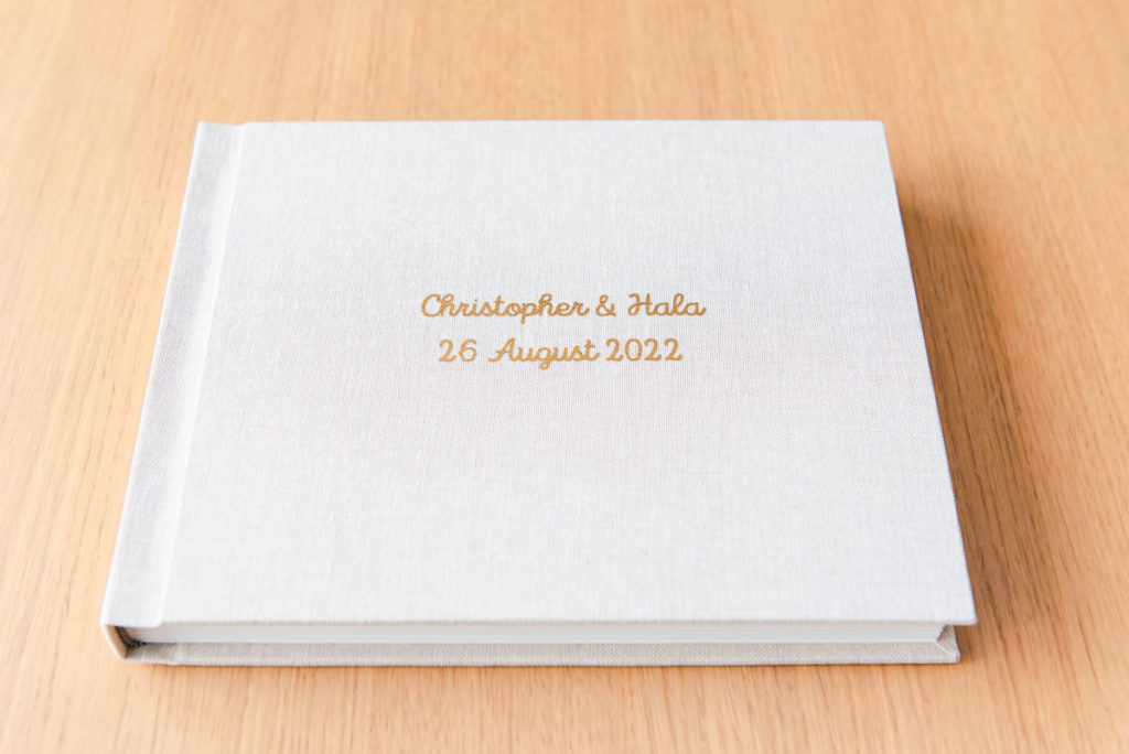 A rectangular wedding album with an oatmeal linen cover and gold lettering on a wooden surface