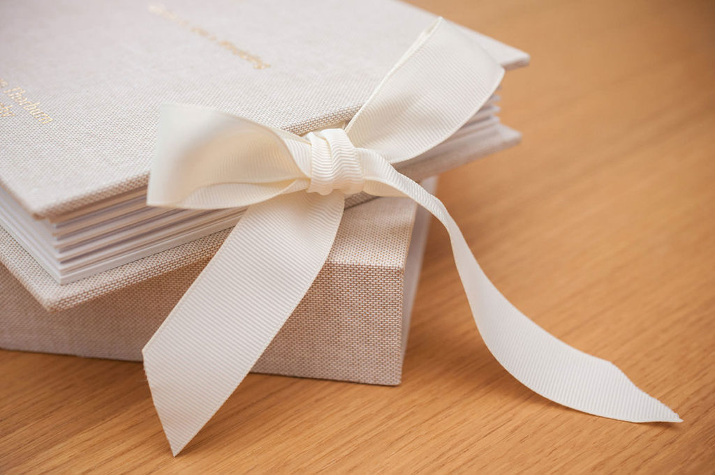 A wedding album with an oatmeal linen cover and gold lettering, tied closed with a cream ribbon tie on a wooden surface