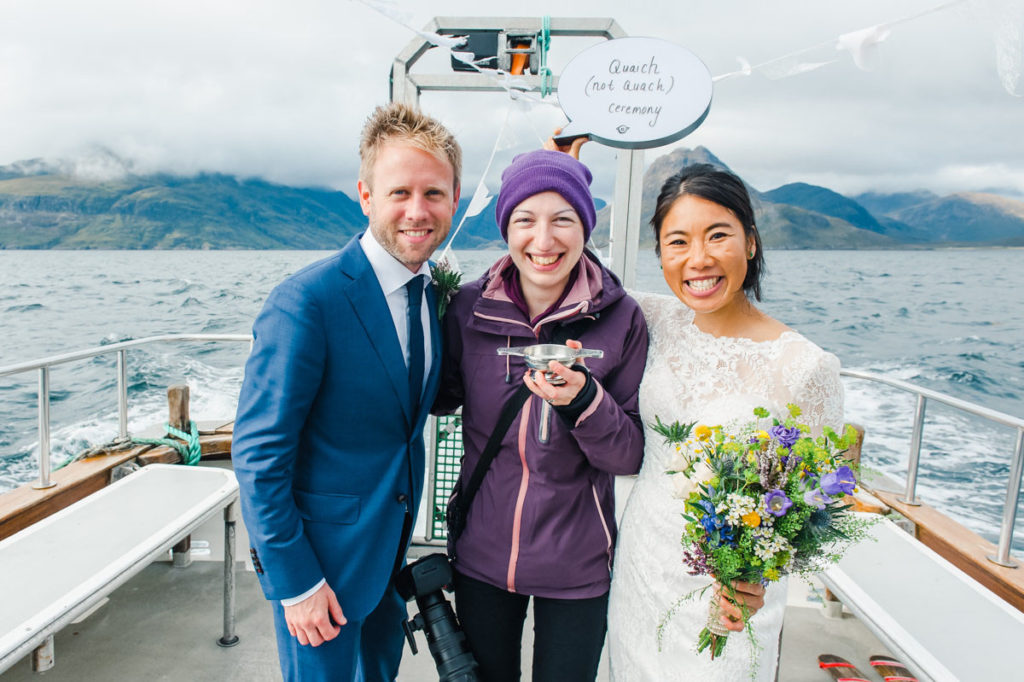 A photographer standing between a bride and groom on the deck of a small boat with a lake and mountains in the background