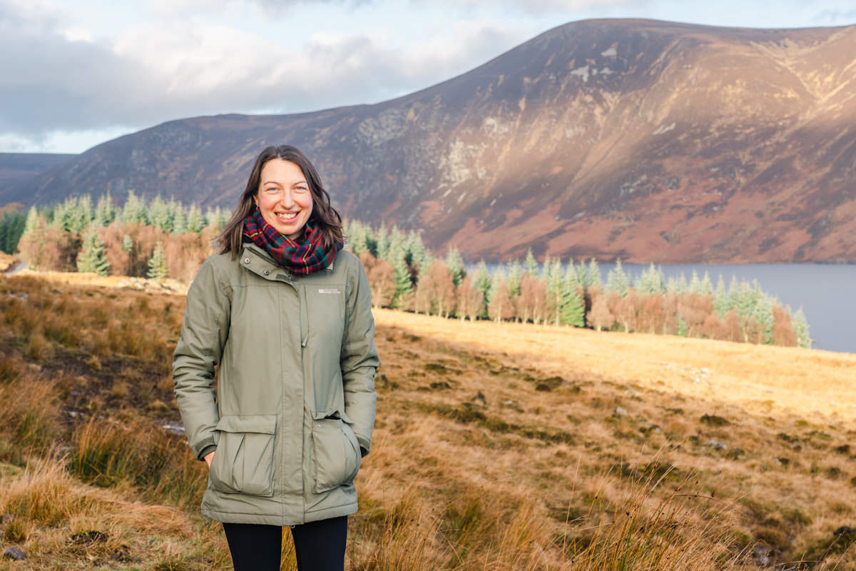 A woman wearing a green jacket and tartan scarf smiling and standing in front of moorland, trees, a loch and a hill