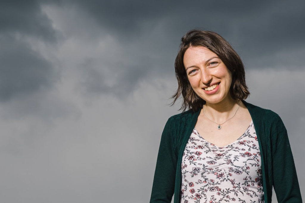 Thirty-year-old white woman wearing a flowery top and green cardigan smiling in front of a dark sky with storm clouds