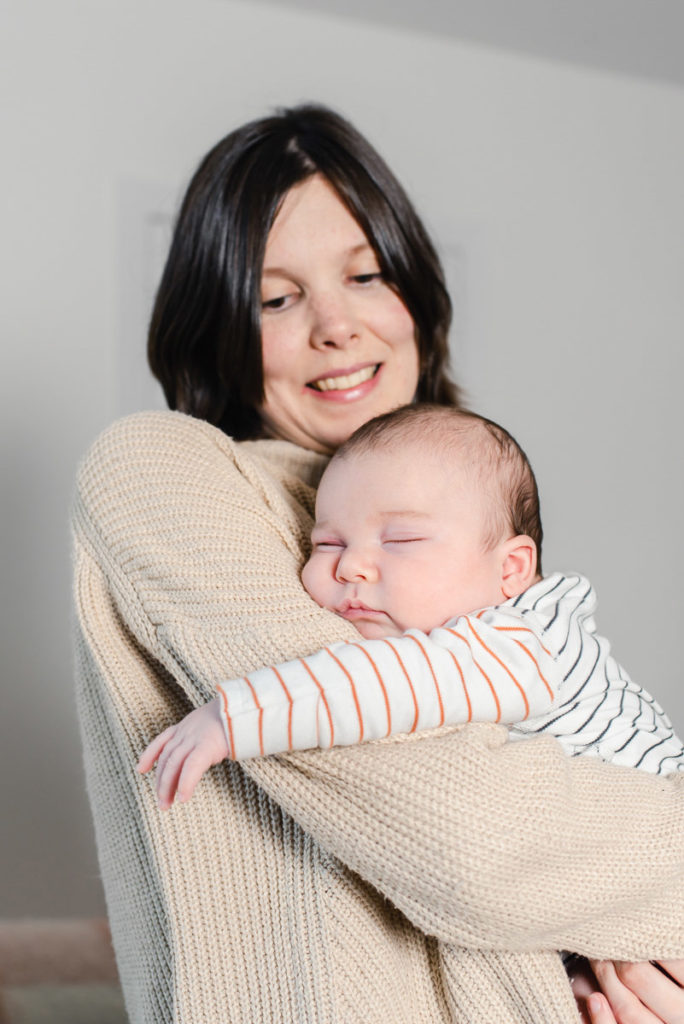 A baby in a striped outfit asleep in the arms of a woman with short brown hair and a beige jumper