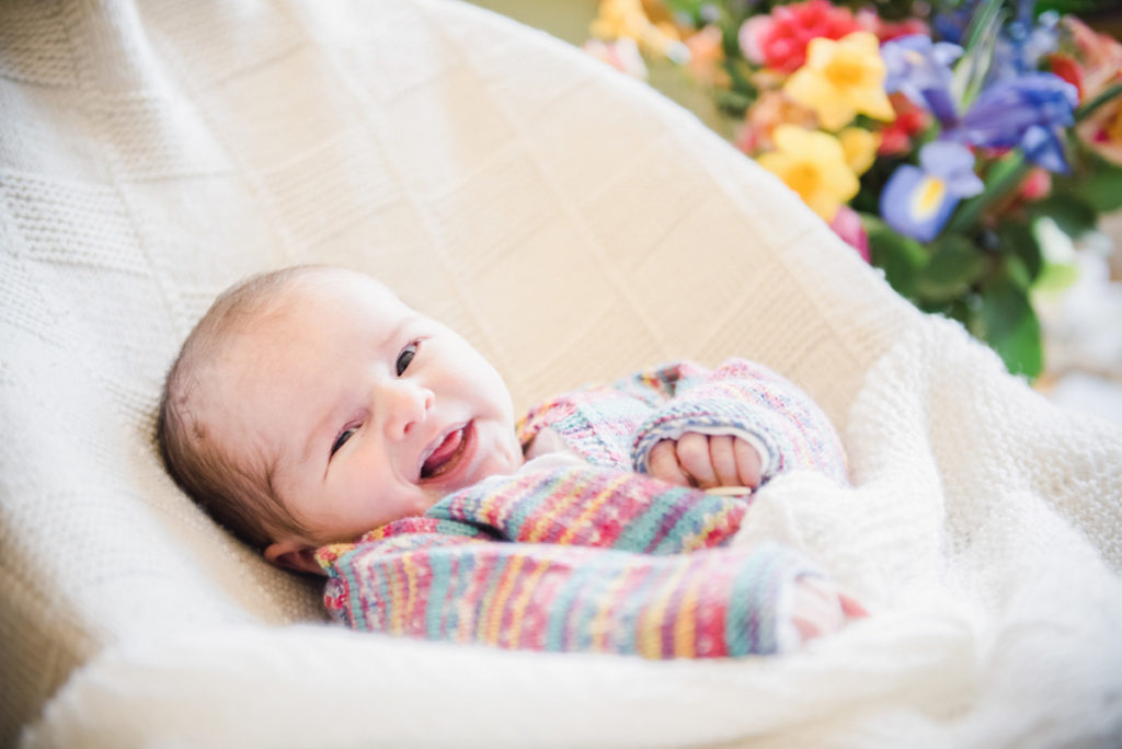 A baby girl in a multi-coloured woollen cardigan laughing while lying in a white bouncer with flowers in the background