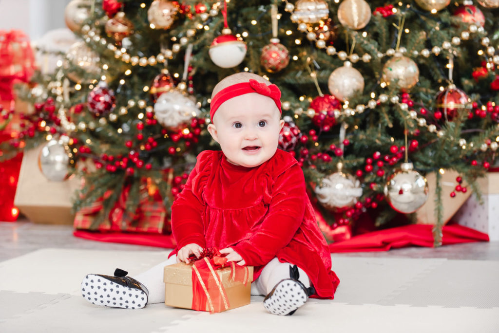 A baby girl in a red dress with a red ribbon on her head sitting with a gold and red parcel in front of a Christmas tree