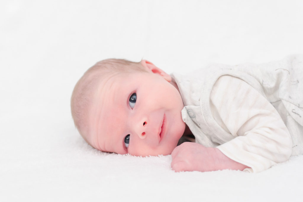 A baby dressed in white lying on its front on a white blanket and gazing upwards