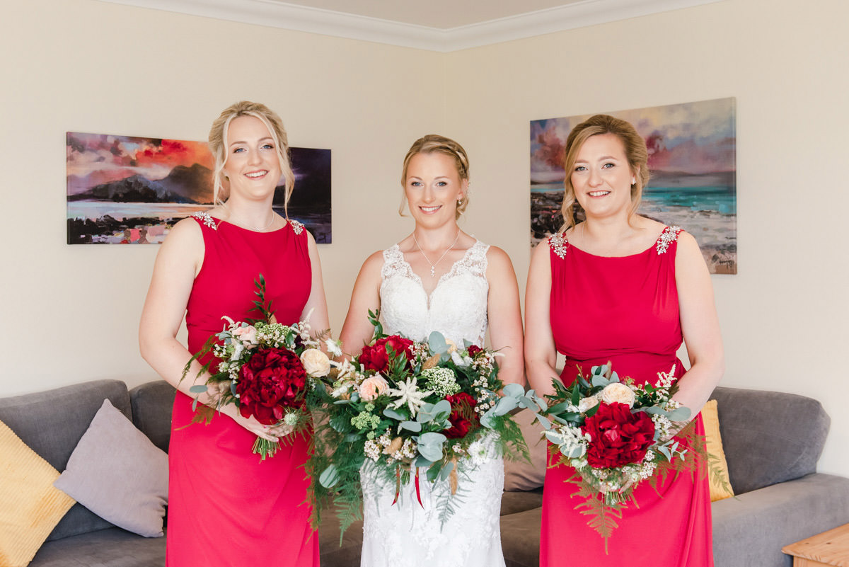 A bride in a white dress standing in between two bridesmaids in red dresses, all holding flowers, in a room with a sofa