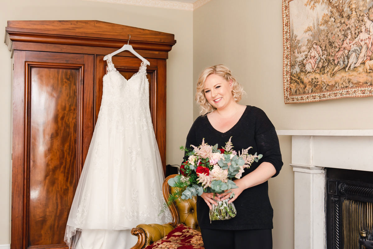 A woman with blonde hair dressed in black holding flowers, standing in front of a white wedding dress hanging on a wardrobe