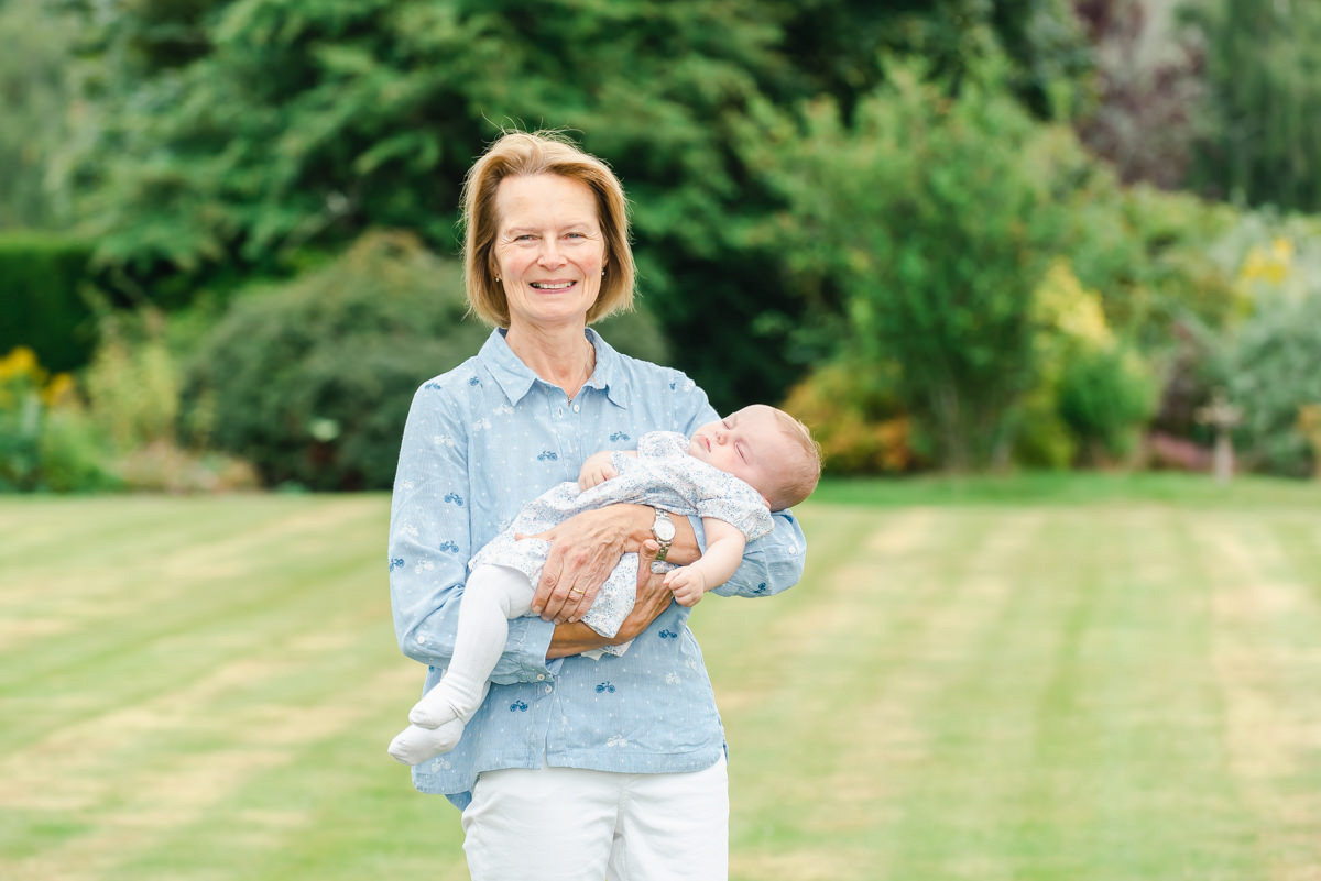 An older woman in a blue shirt and white trousers holding a sleeping baby girl with a lawn and trees in the background