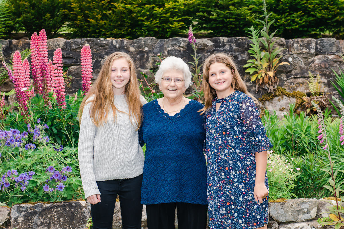 A woman with white hair and a navy top standing between two teenage girls in front of flowers and a stone wall