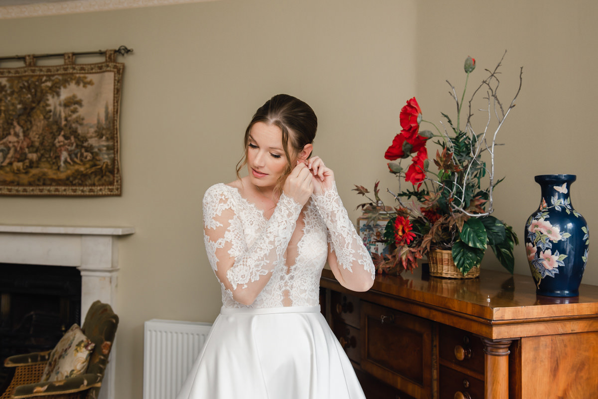 A bride in a white dress with lace sleeves looking down while inserting an earring in a room with a wooden chest and flowers