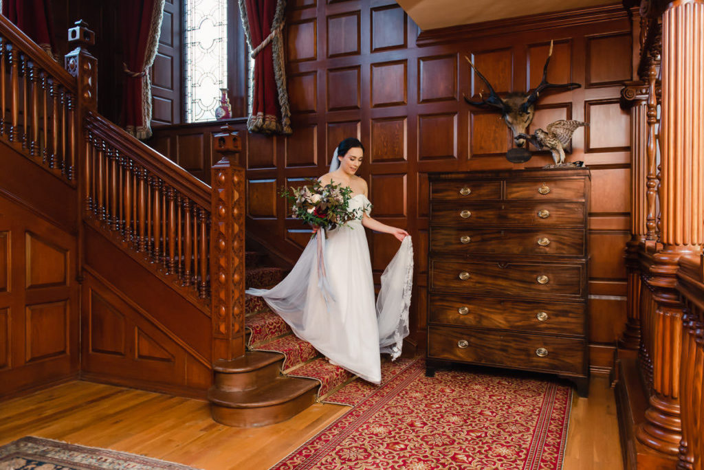 An elegant bride in a white wedding dress holding her flowers and veil while descending a wooden staircase