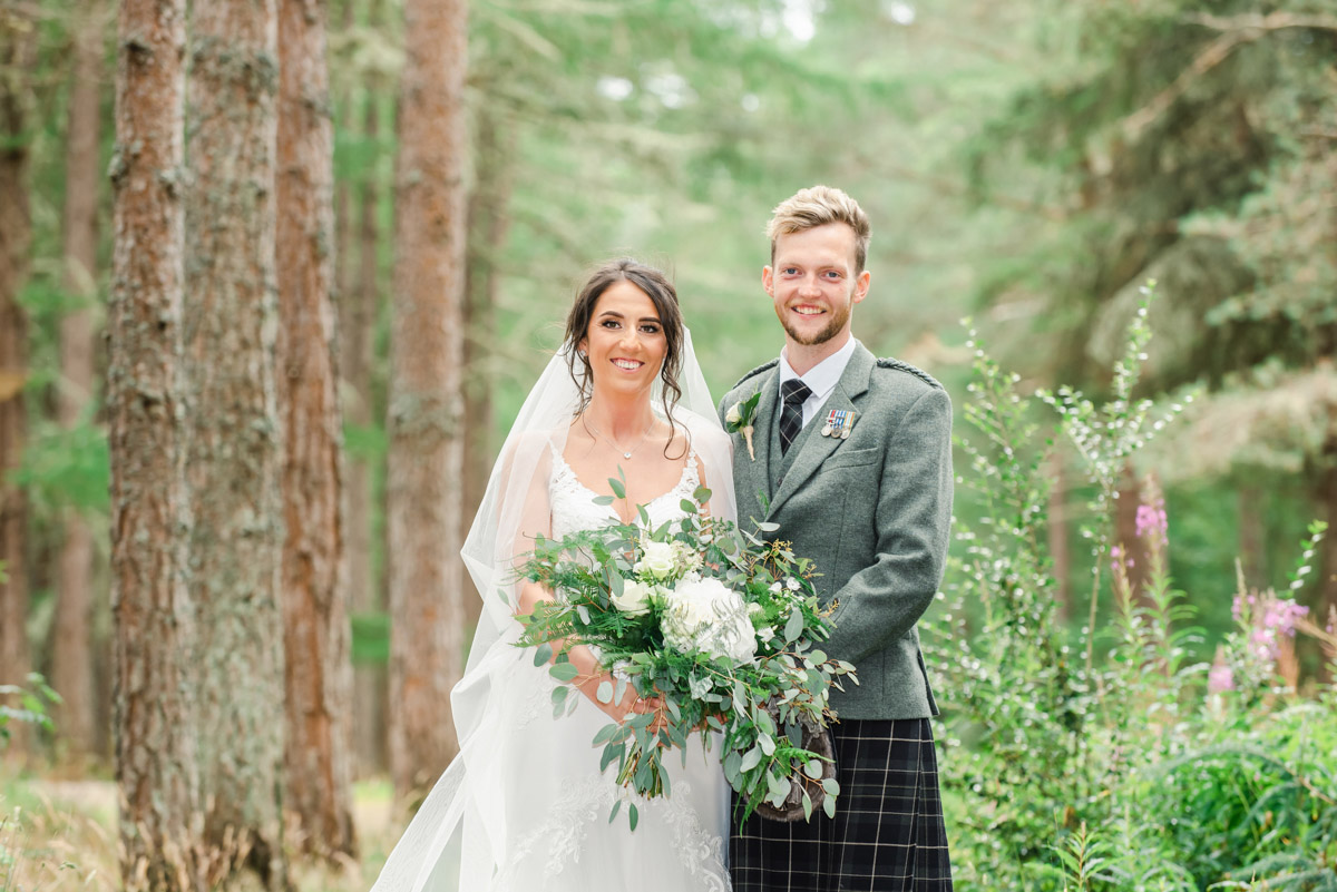 A bride in a white dress and a groom in a kilt and green jacket standing beside trees and flowers in Daviot Woods