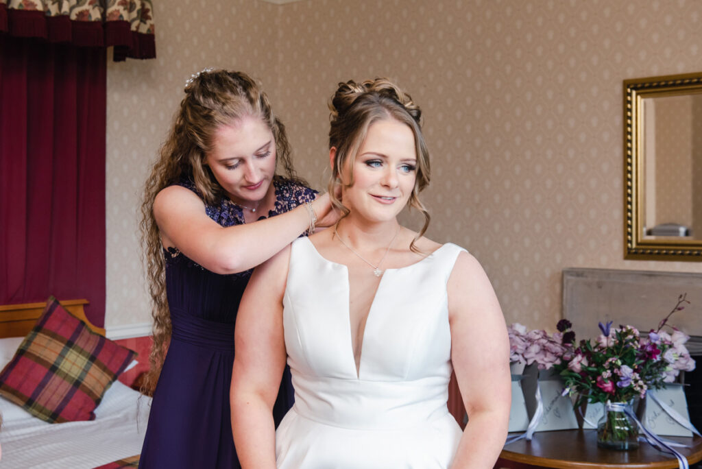 A woman helping a bride with her necklace in a bedroom at the Kincraig Castle Hotel near Invergordon