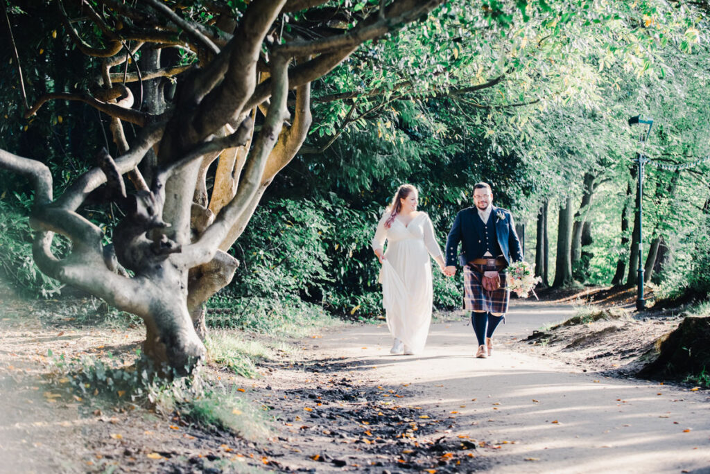 A bride and groom holding hands and walking on a path in the woods at Ness Islands in Inverness