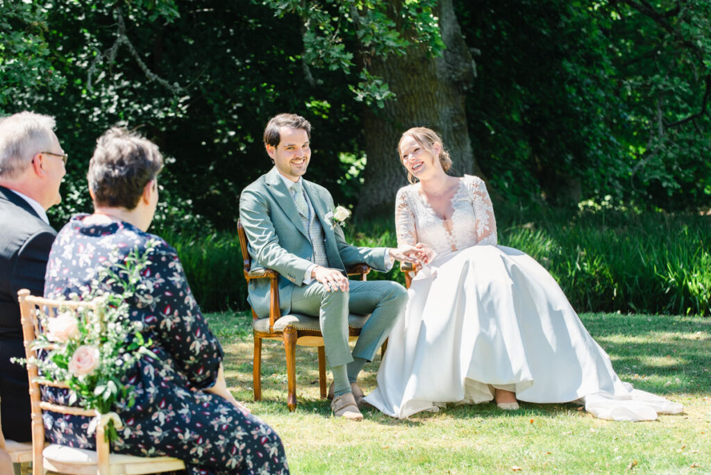 A bride and groom sitting and holding hands during their wedding ceremony on the lawn at Pitcalzean House
