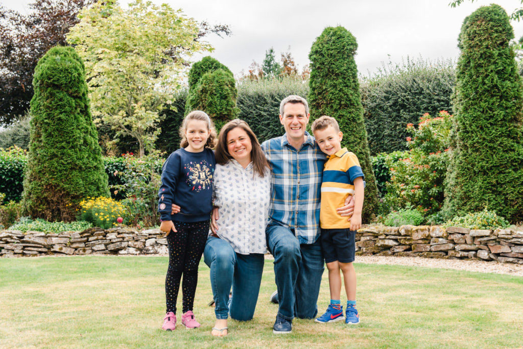 A family of four with the parents and their young daughter and son on grass in a garden in front of a stone wall and hedges