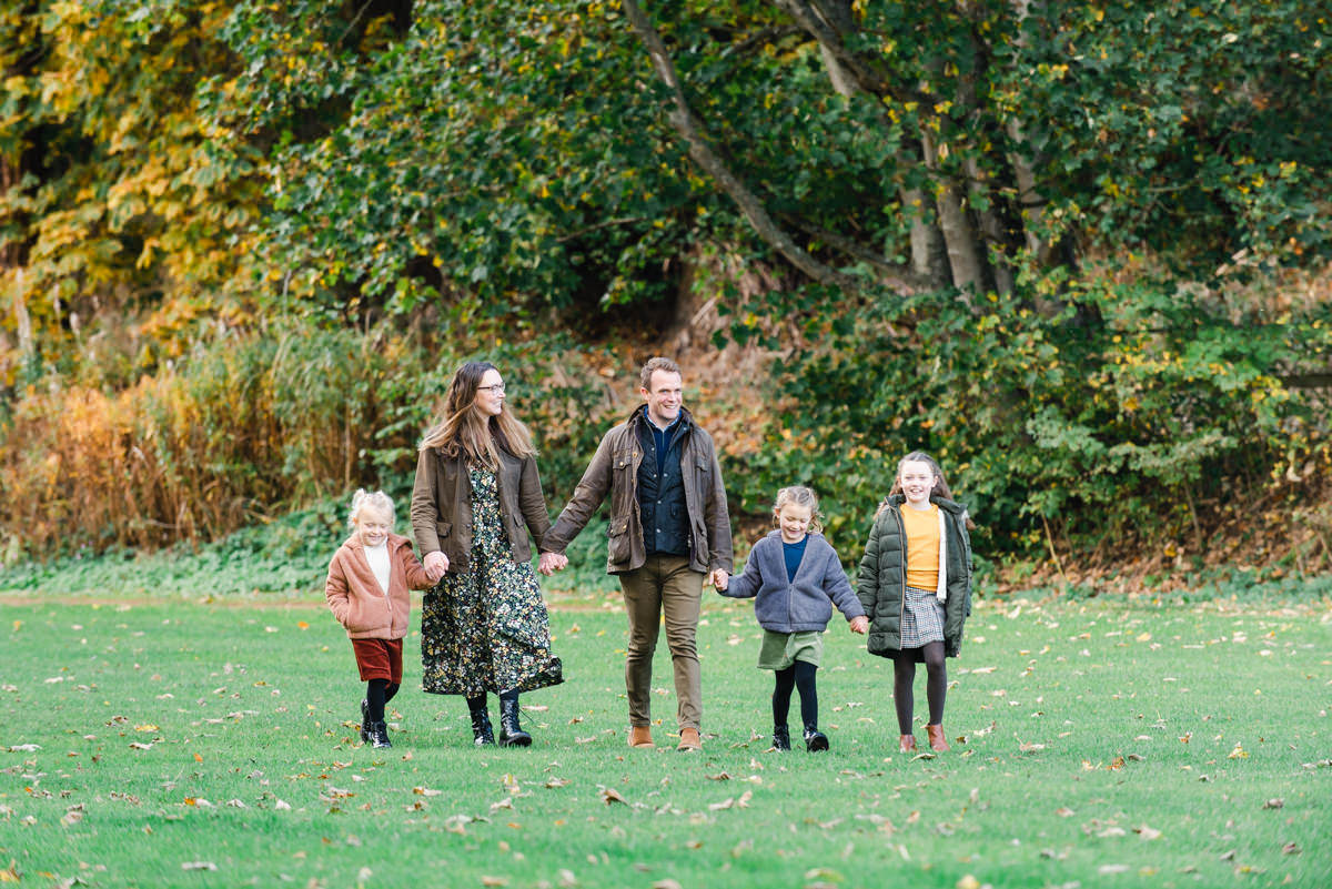 Family of five with parents and their three young daughters holding hands and walking across grass in front of trees