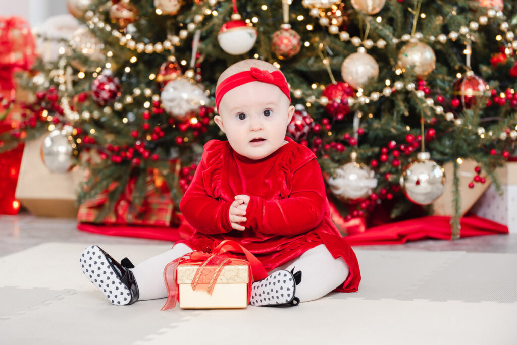 An infant in a red dress sits in front of a Christmas tree with her present during an Inverness Family Photo Shoot
