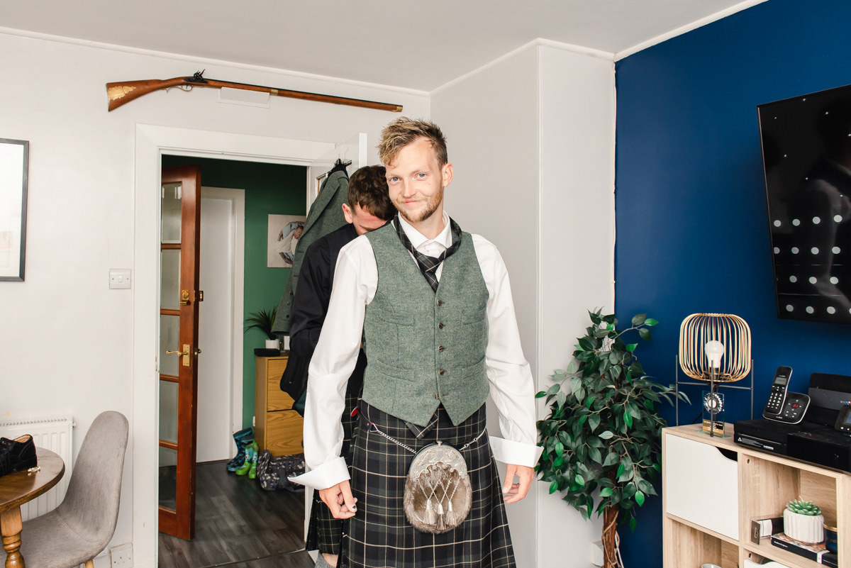 A man in a kilt and waistcoat being helped by another man in a living room with a gun above the door