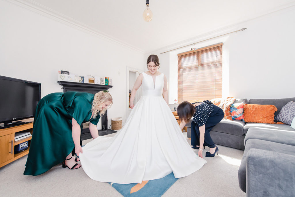 A bridesmaid in a green dress and a woman in a navy outfit fanning a white wedding dress worn by a bride in a neutral lounge