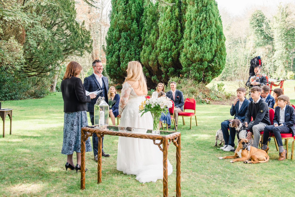 Bride and groom face each other in garden ceremony with seated guests and piper looking on at Bunchrew house near Inverness