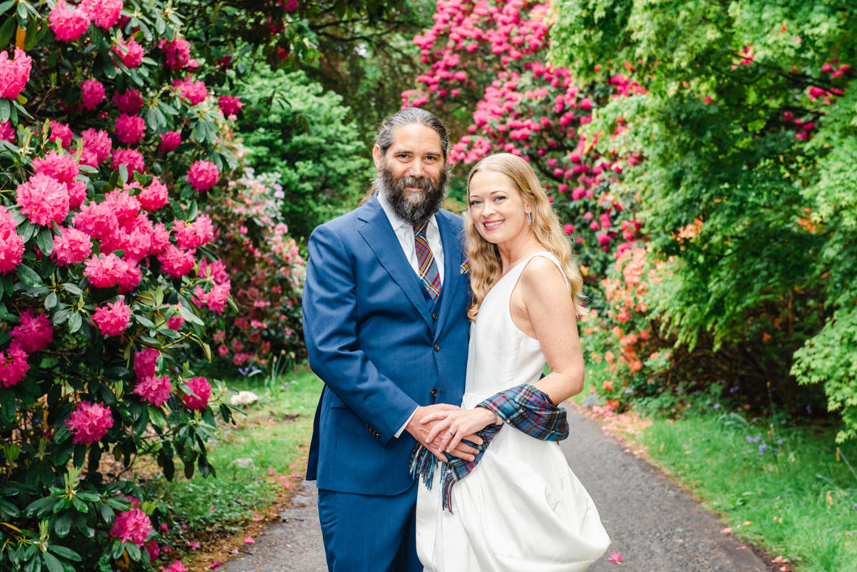 A groom in a blue suite holds his bride while standing on a path surrounded by rhododendrons with red flowers in Burgie House