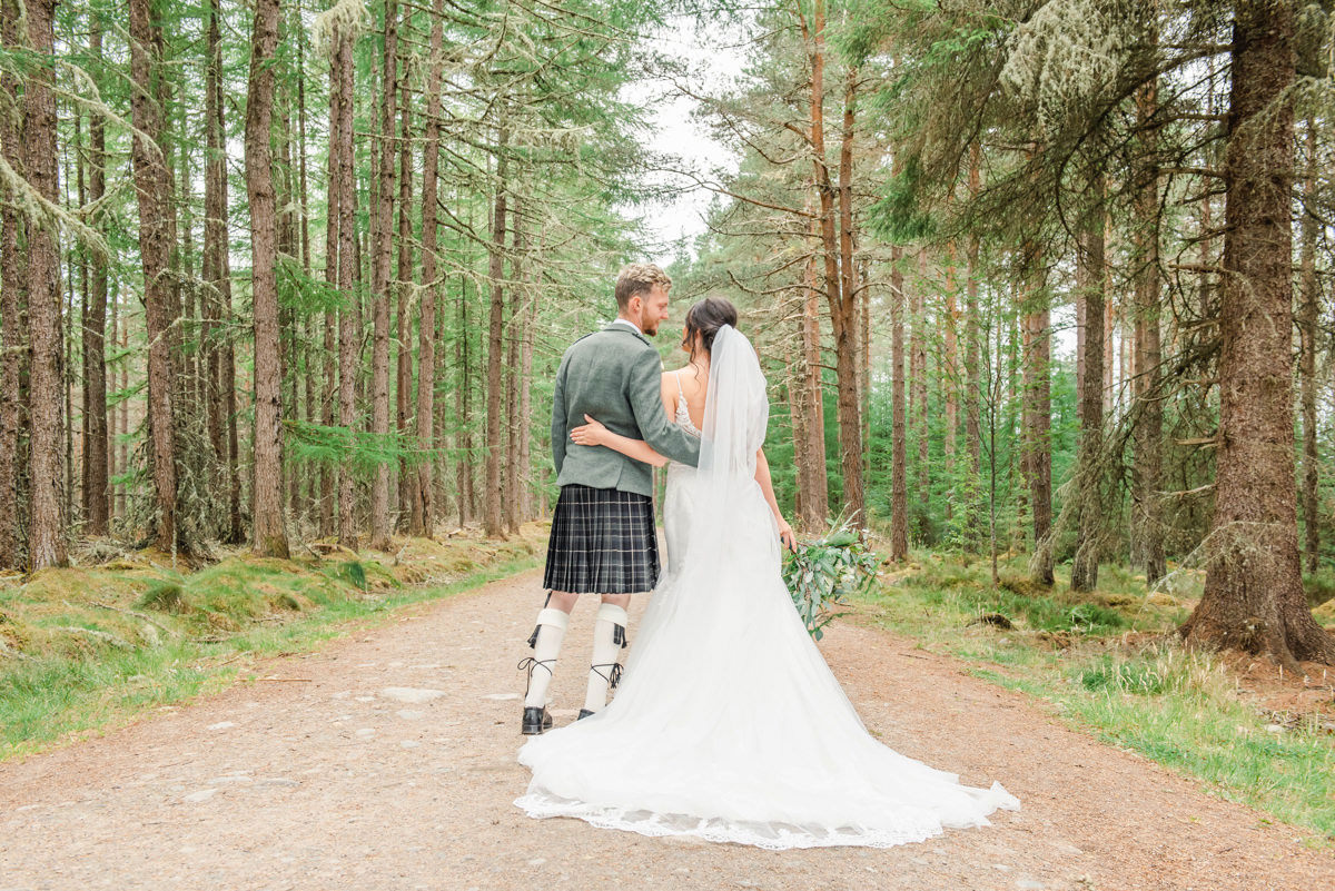 A groom in a green tweed jacket stands with his bride backs to camera looking along a woodland path in Daviot woods, Inverness