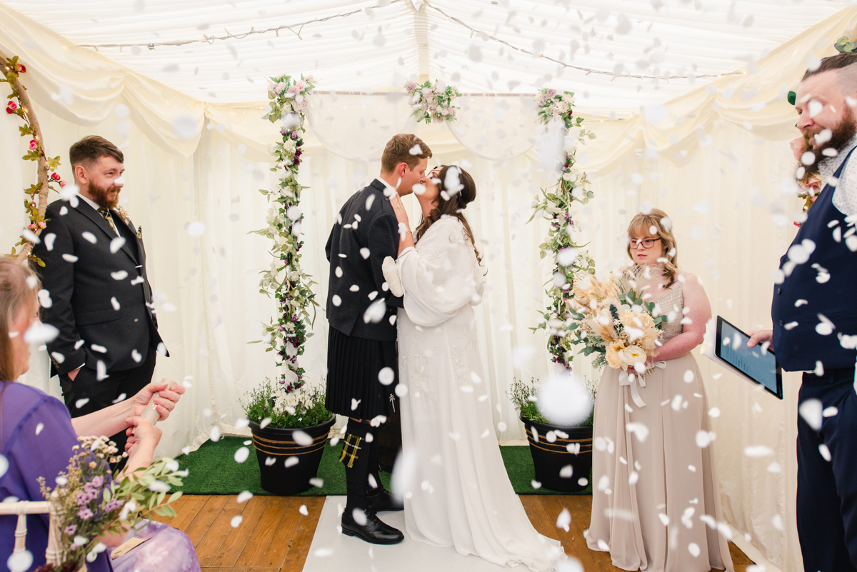 Bride and groom kiss while white petals fall from above and guest look on in a marquee wedding in Dallas near Forres
