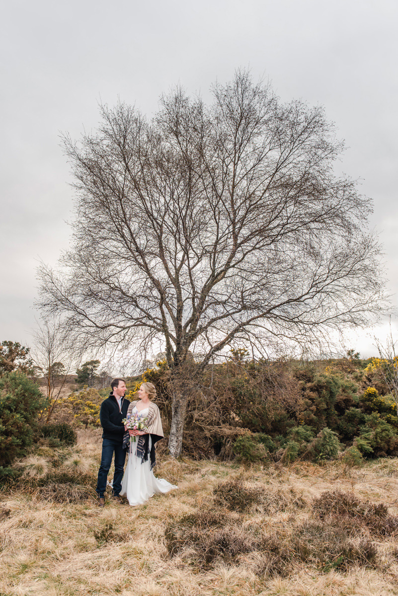 A groom in jeans stands next to a bride in a cream shawl holding a bouquet in front of a birch tree in Glen Convinth