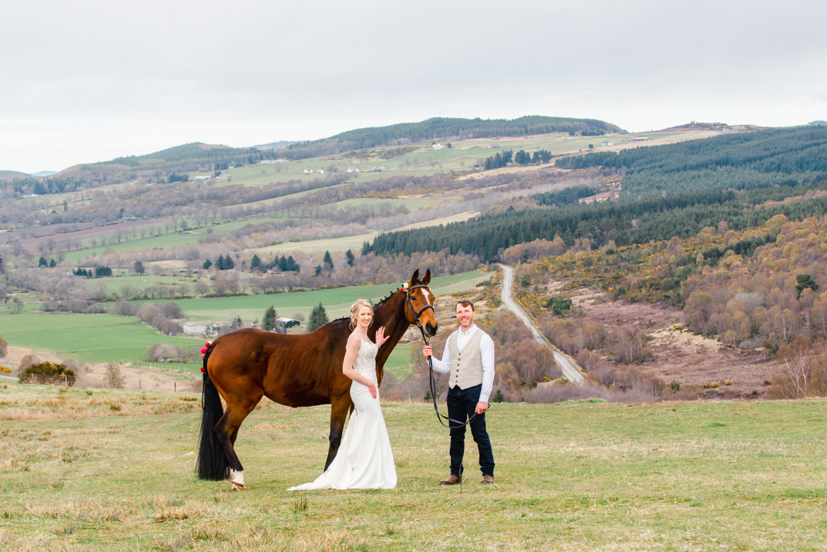 A bride and groom stand next top a large brown horse in a field overlooking hills on Glen Convinth estate