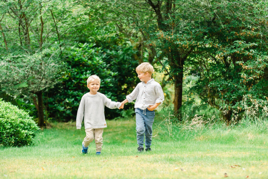 A boy and his younger brother hold hands while walking though a grassy woodland during a Highland Family Photo Shoot
