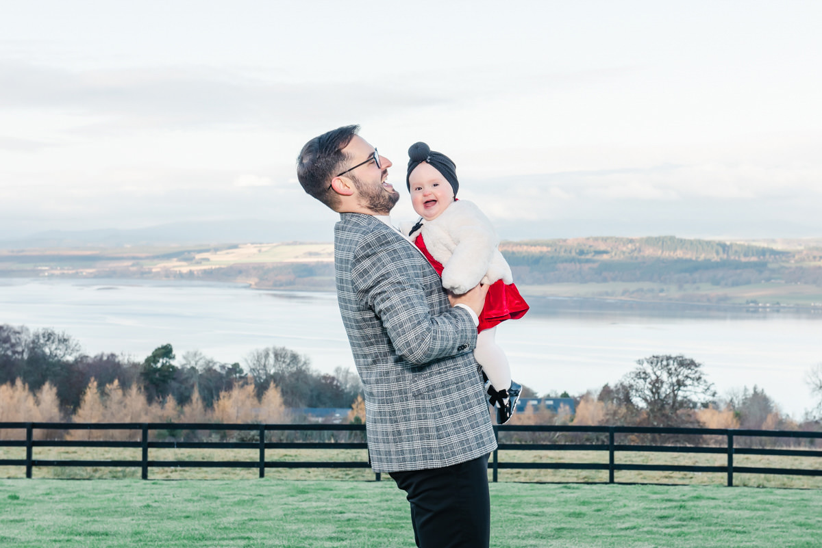 A father in a grey jackets holds his smiling baby daughter in a red dress to his chest during an outdoors Family Photo shoot