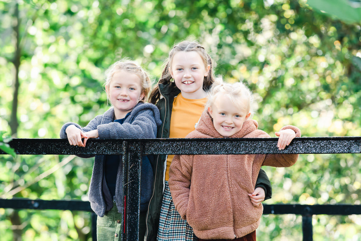 Three young sisters in winter jackets stand on a wooden bridge in a woodland setting during a family Photo Shoot at Rosemarkie